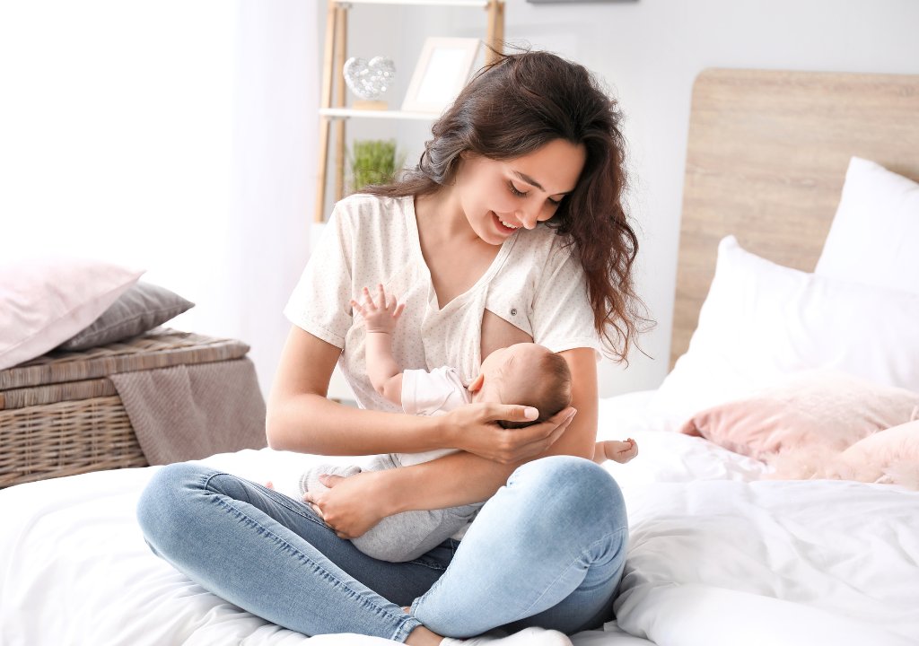 Phụ nữ đang cho con bú bế em bé và nhìn con trong không gian phòng ngủ sáng sủa