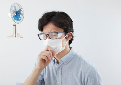 Người đàn ông đeo khẩu trang, ho và dùng khăn giấy trong phòng có quạt đang thổi, biểu hiện kích ứng cổ họng khi nằm quạt.